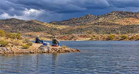 Two anglers fishing from the rocky shoreline of Alamo Lake with fishing gear and storm clouds over the desert mountains