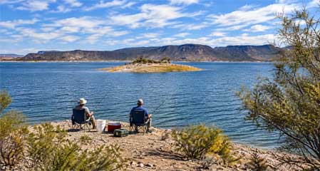 Two anglers fishing from the shoreline of Lake Pleasant Arizona with a bait bucket, cooler and tackle box beside the water